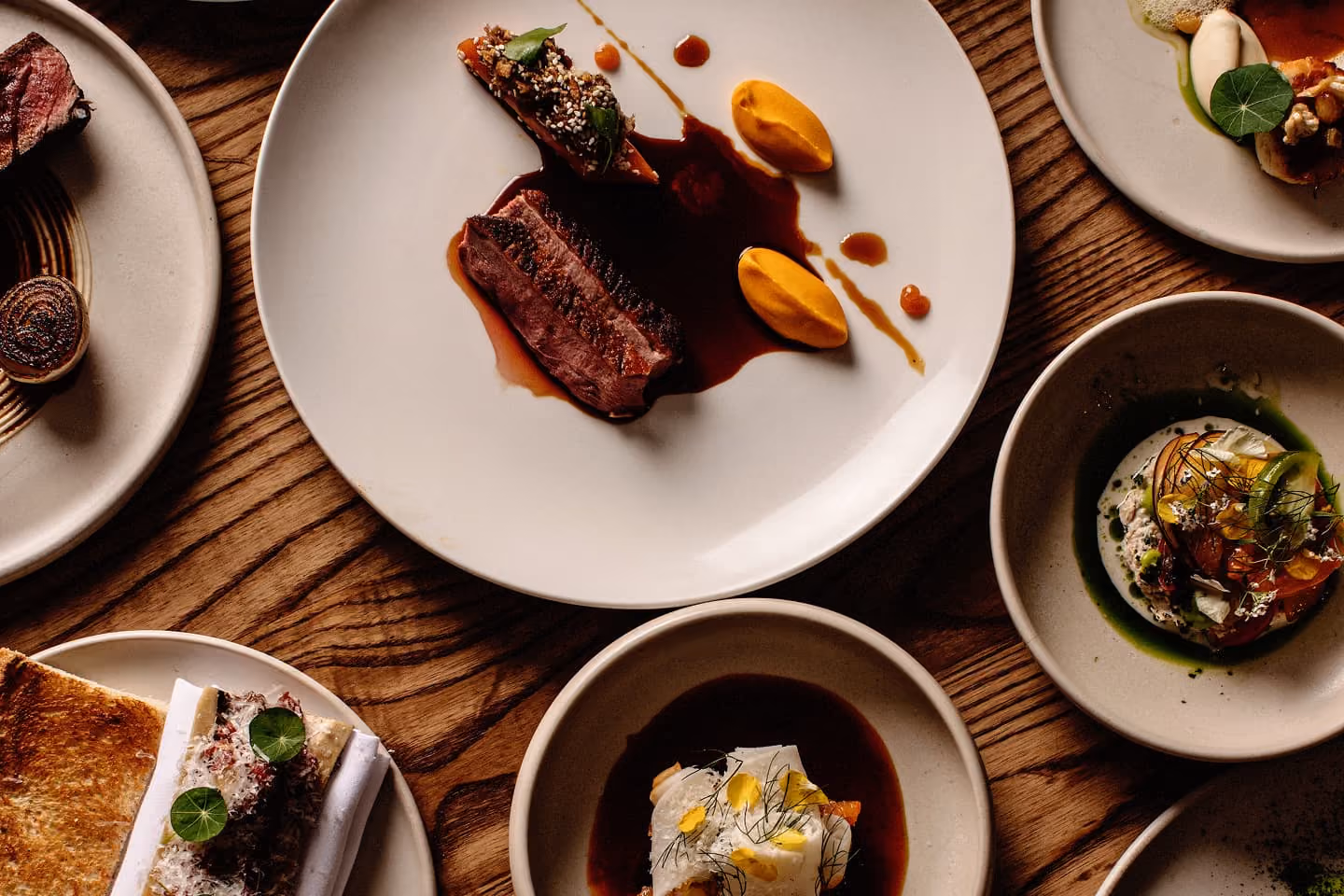 Elegant gourmet dishes on ceramic plates set on a wooden table, featuring meats, sauces, and garnishes.
