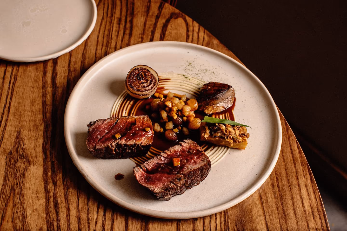 Plate with seared steak slices, grilled onion, sautéed mushrooms, and a garnish on a wooden table.