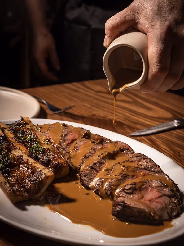 Hand pouring brown gravy over sliced steak and grilled bone marrow on a white plate.
