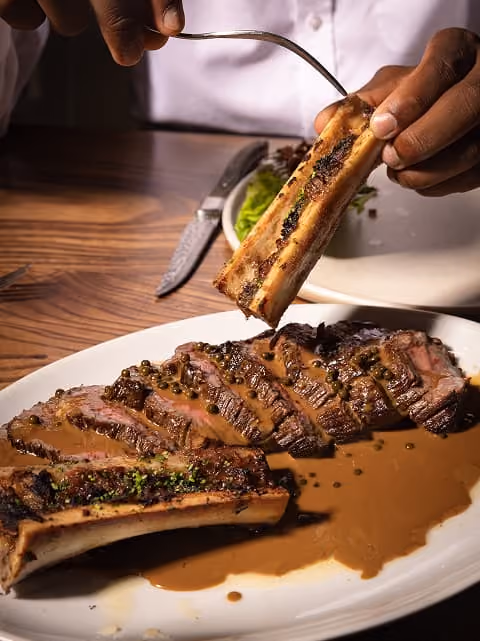 Hand holding a rib bone above sliced steak with sauce on a white plate.