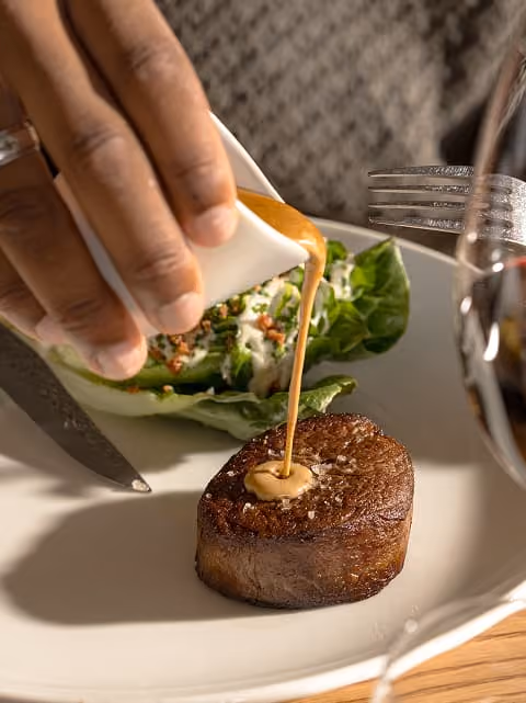 Hand pouring sauce onto a cooked filet mignon steak on a plate with salad and utensils nearby.