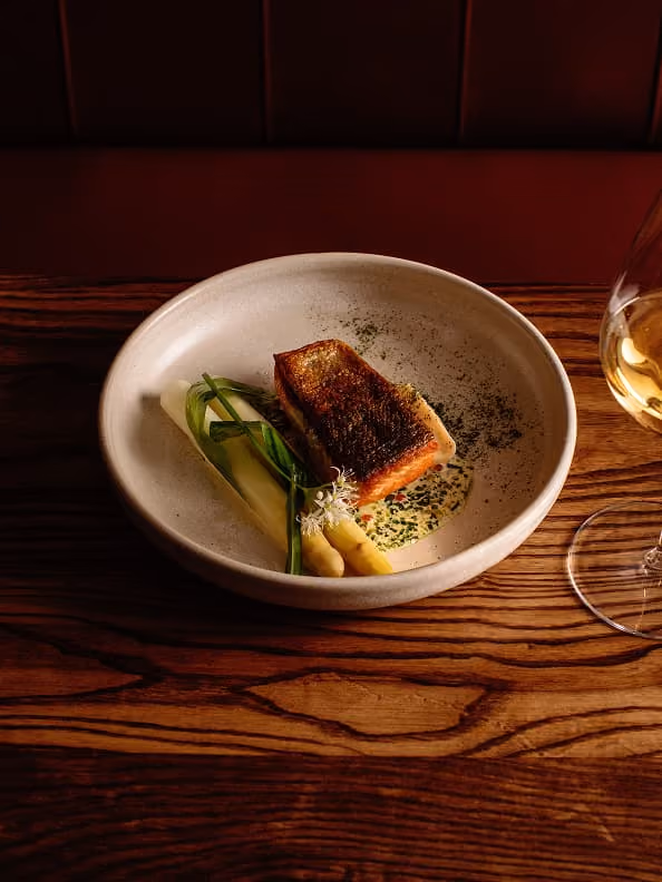Seared fish fillet with white asparagus and green garnish on a sauce in a bowl with a glass of white wine nearby.