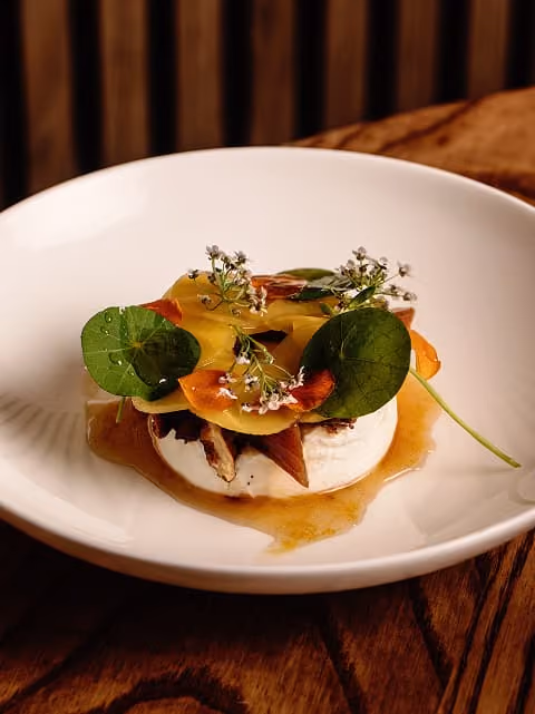 Gourmet dish with cheese, edible flowers, leaves, and sauce on a white plate on a wooden table.