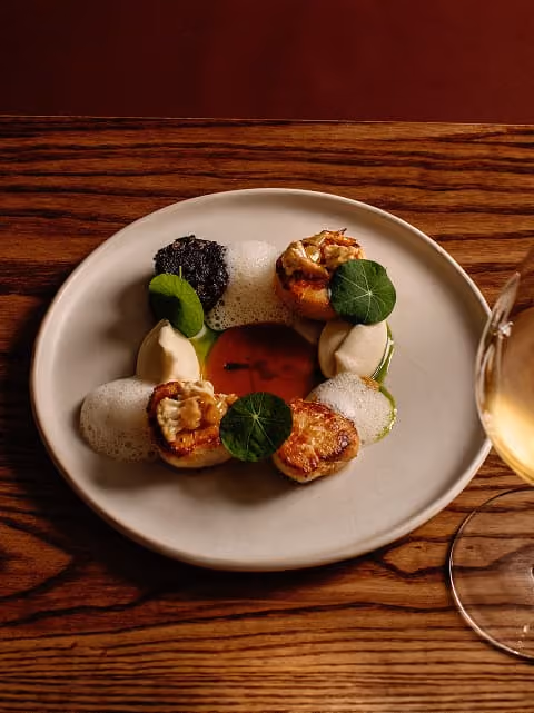 Gourmet dish with three seared scallops, green leaves, sauce, and foam on a white plate with a wine glass.