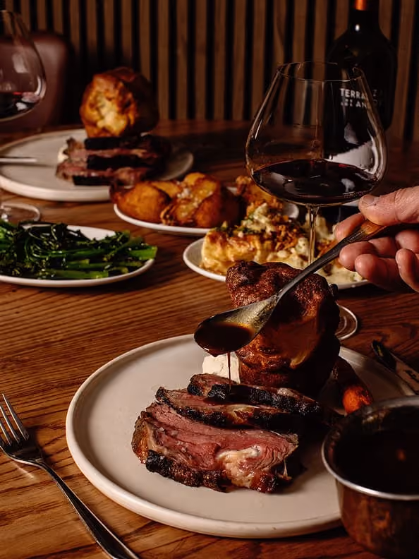 Plate with slices of roast beef and Yorkshire pudding, hand pouring gravy, wine glass and side dishes on table.