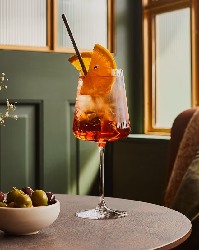 Orange spritz garnished with a slice of orange, on a table beside a bowl of olives