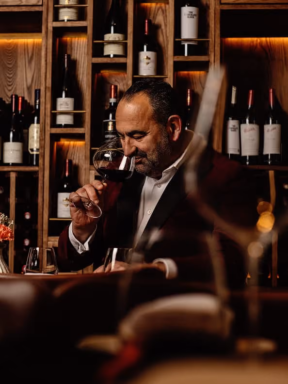 Man in suit smelling red wine in glass with wine bottles on wooden shelves behind him.