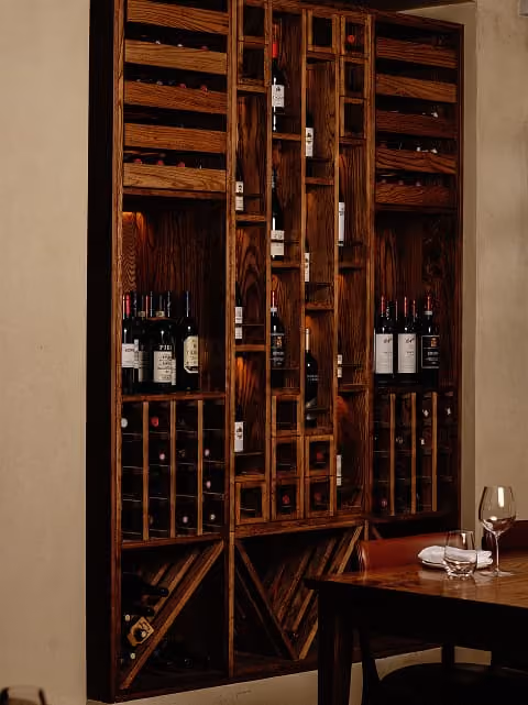 Wooden wine rack filled with various wine bottles beside a table with a wine glass and folded napkins.