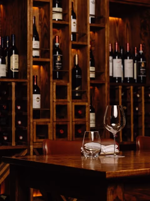 Wine bottles displayed in wooden shelves behind a table with empty wine and water glasses.