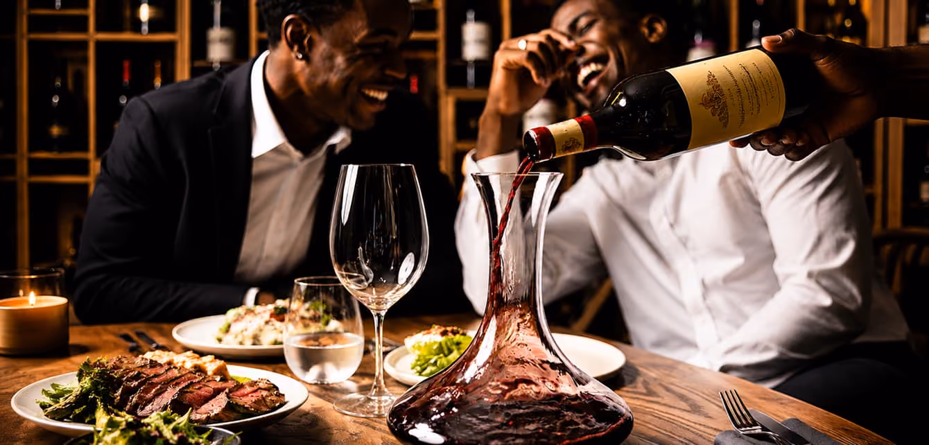 Two men smiling at a table as red wine is poured into a decanter with plated food nearby.