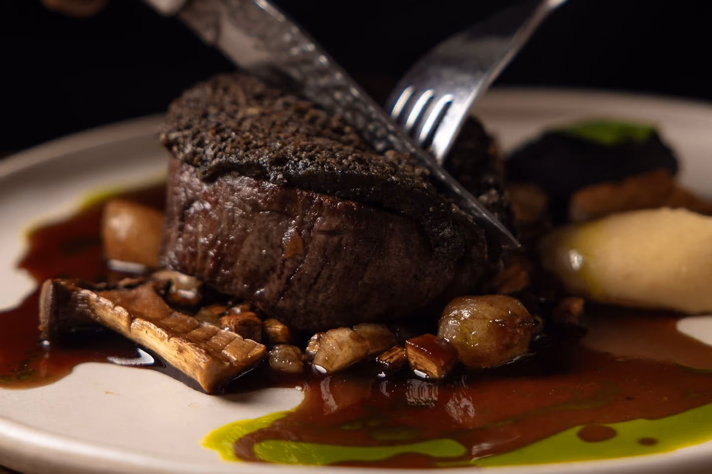 Fork and knife cutting into a seared steak served with mushrooms and sauce on a white plate.