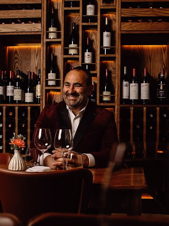 Smiling man in dark jacket sitting at a table in front of a wooden wine rack with bottles.