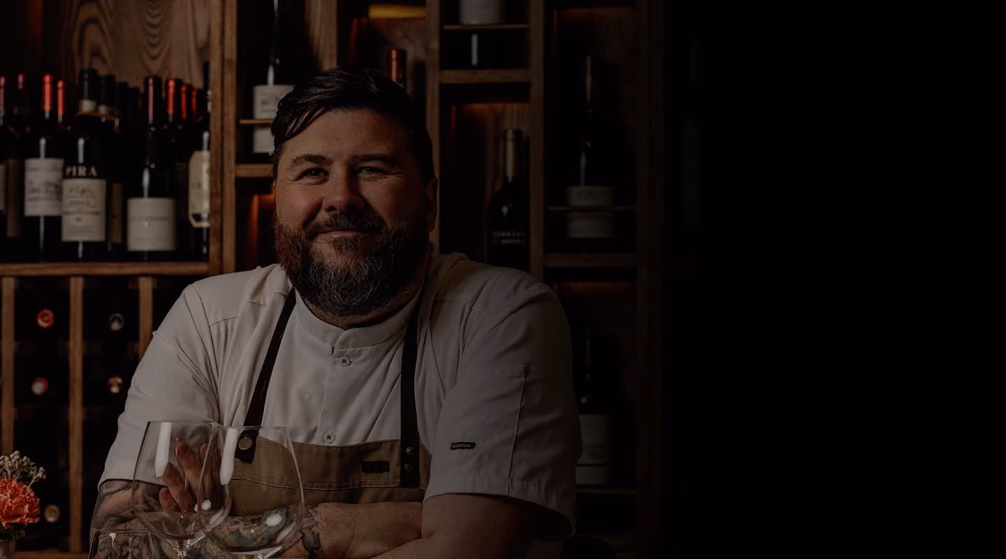 Bearded chef in white uniform and apron smiling with folded arms at a wine cellar bar.