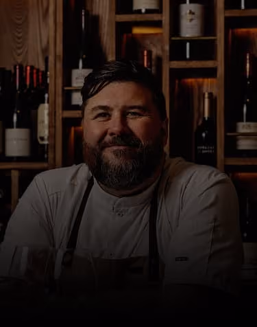 Smiling male chef with beard in a kitchen with wooden shelves and wine bottles.