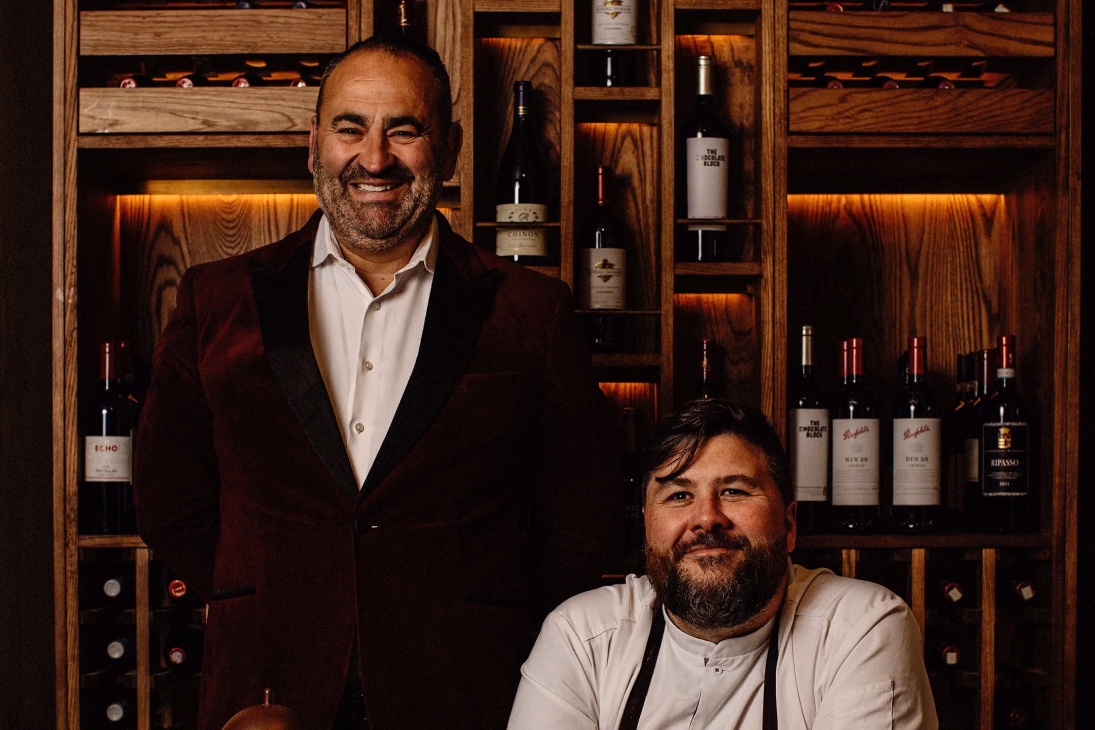 Two men smiling in front of a wooden wine rack filled with bottles in warm lighting.