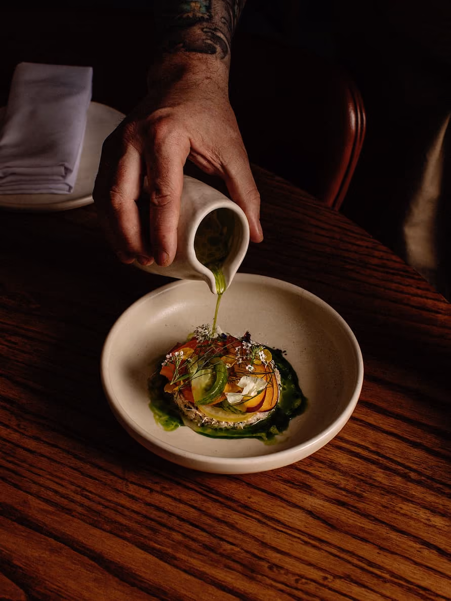 A person pours green sauce from a small pitcher onto a beautifully plated gourmet dish decorated with fresh herbs and edible flowers.