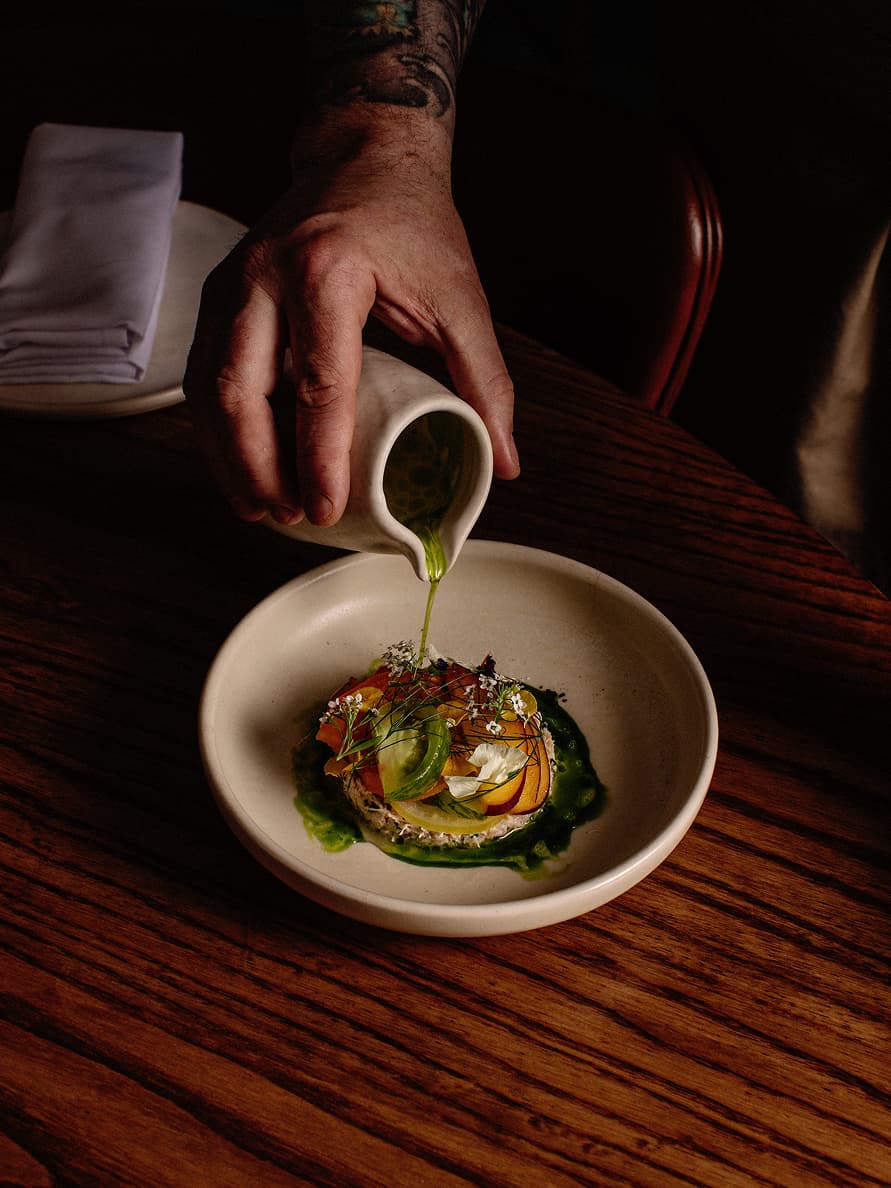 A person pours green sauce from a small pitcher onto a beautifully plated gourmet dish decorated with fresh herbs and edible flowers.