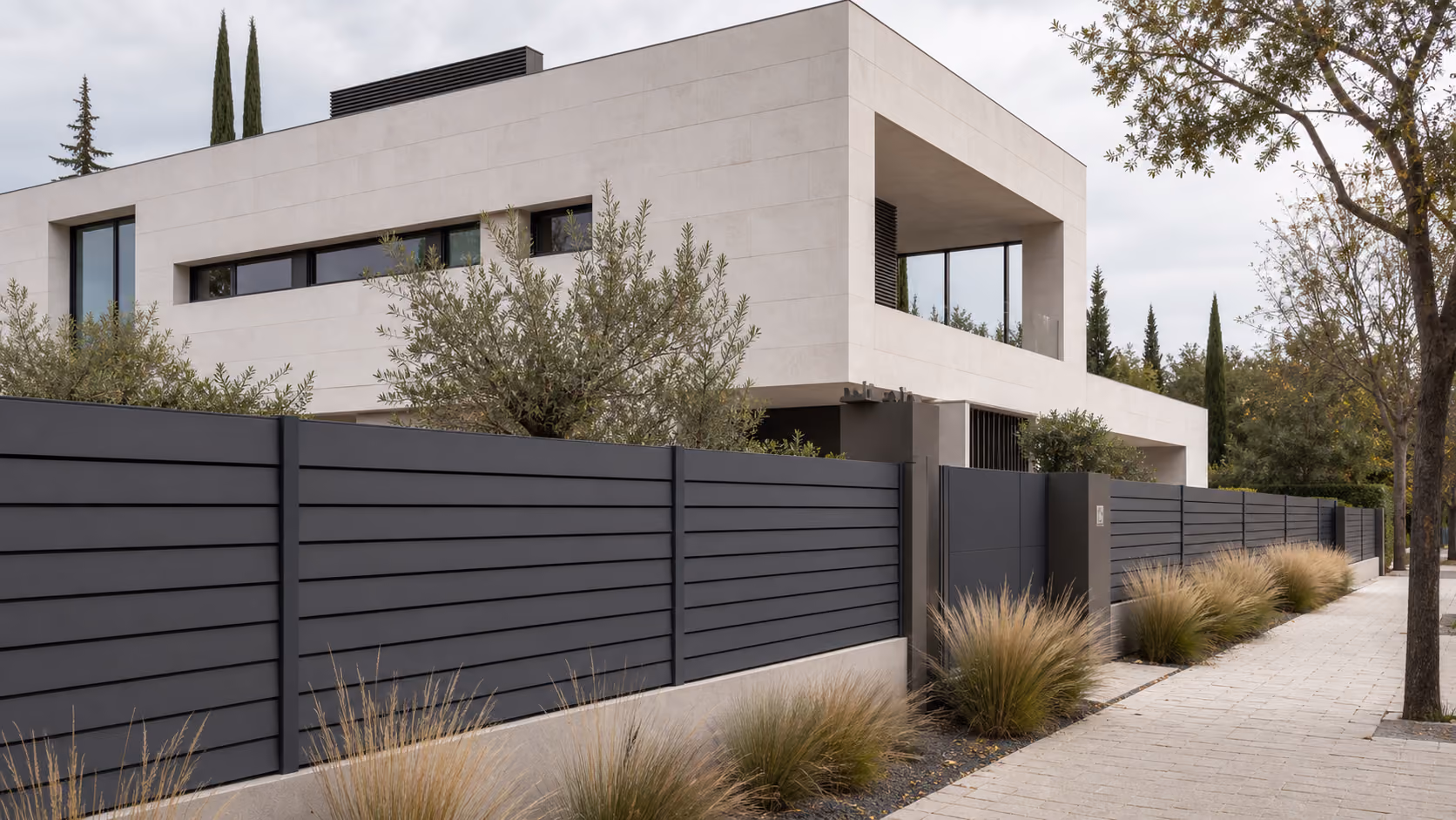 Modern house with beige walls behind a long, dark gray horizontal slat fence and ornamental grasses.