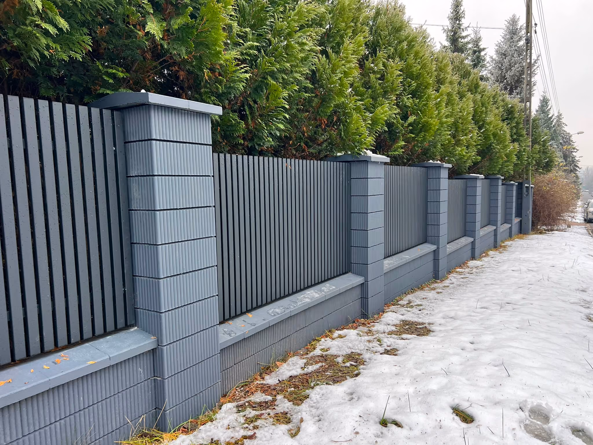 Gray fence with vertical slats and pillars along a snowy sidewalk next to green trees.