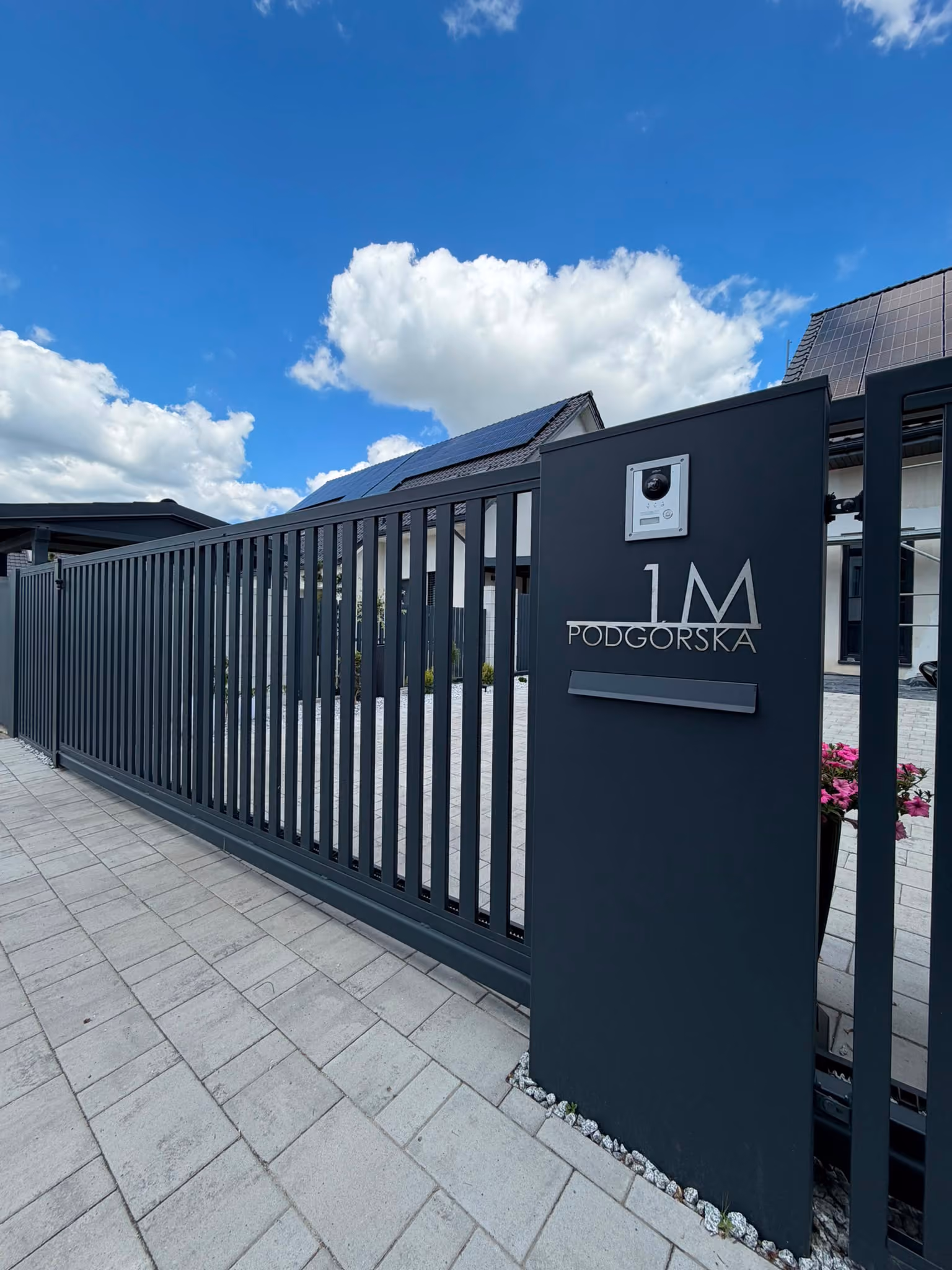 Modern dark metal gate with house number 1M Podgorska and solar panels on the roof behind it.