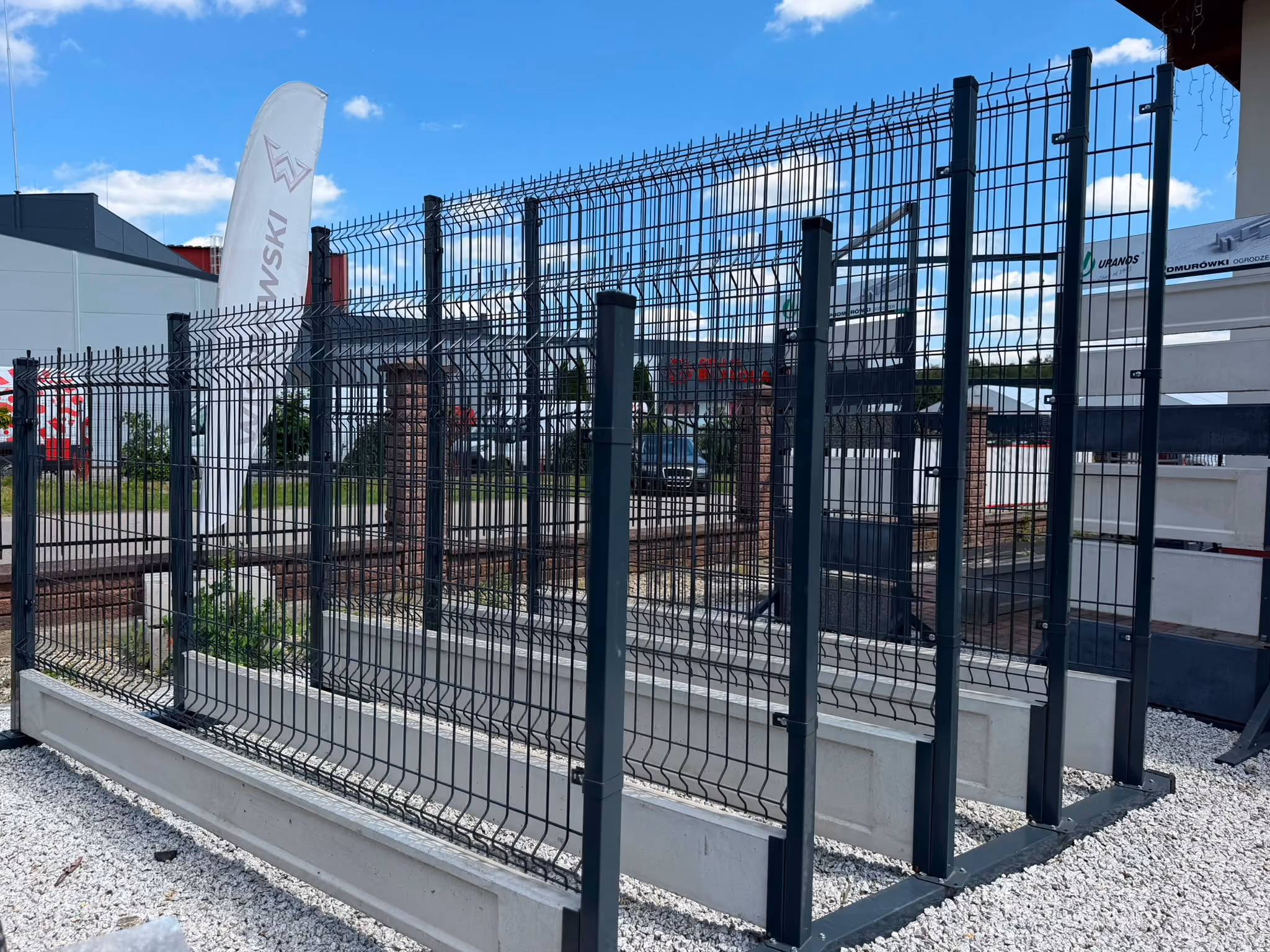 Black metal wire mesh fence panels mounted on concrete bases outdoors under blue sky.