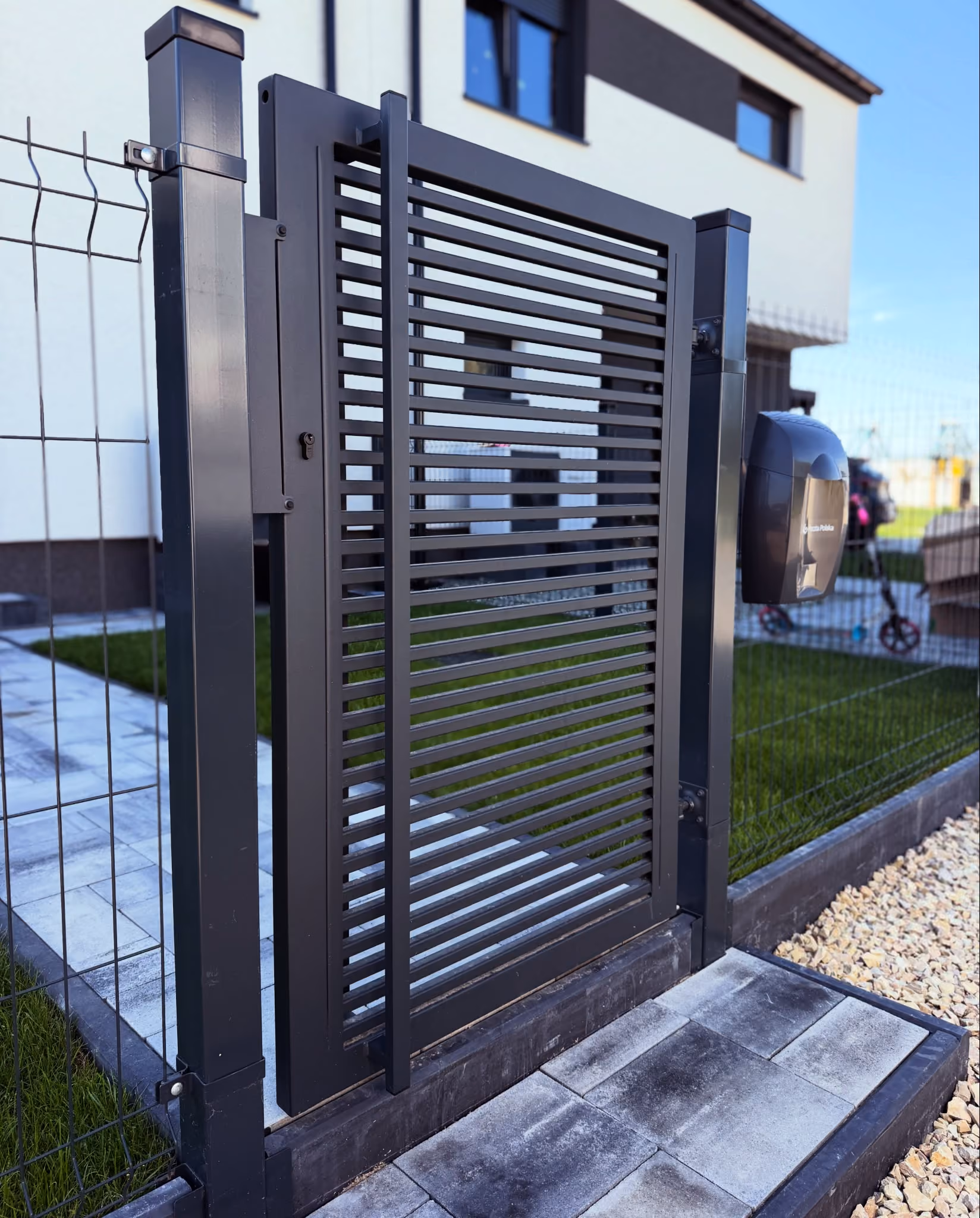 Modern black metal gate with horizontal slats attached to a fence in front of a house.