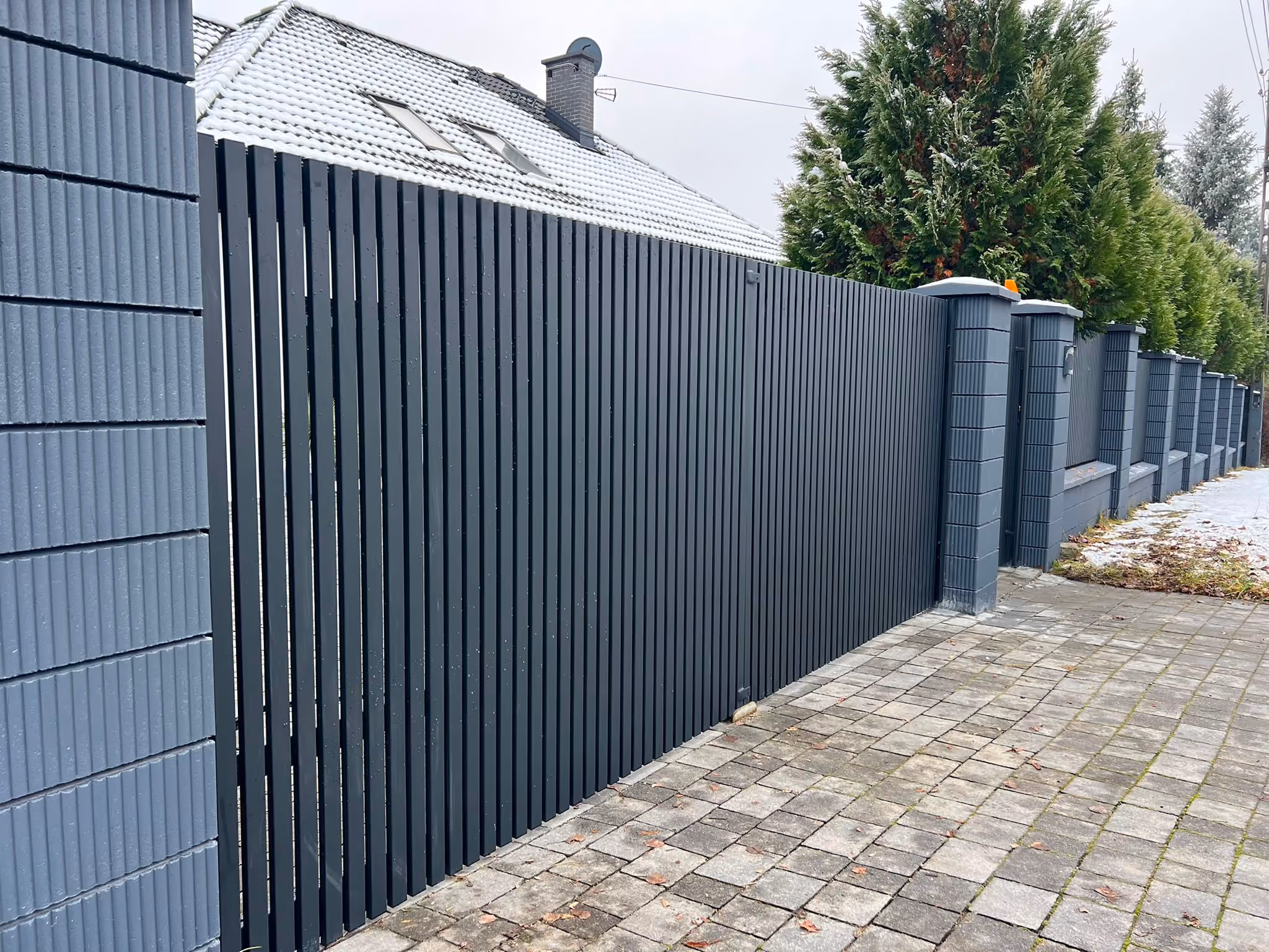 Tall dark gray vertical slat fence with pillars lining a stone-paved sidewalk beside a snowy roof house.