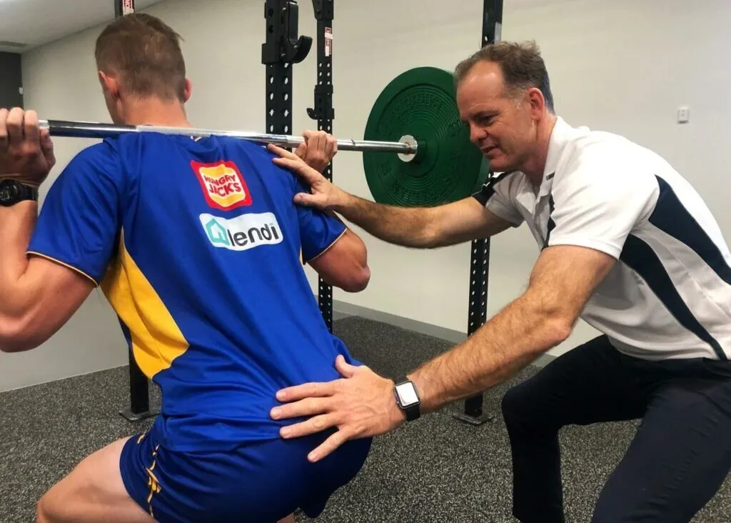 Physiotherapist coaching an athlete through a barbell squat during sports rehabilitation