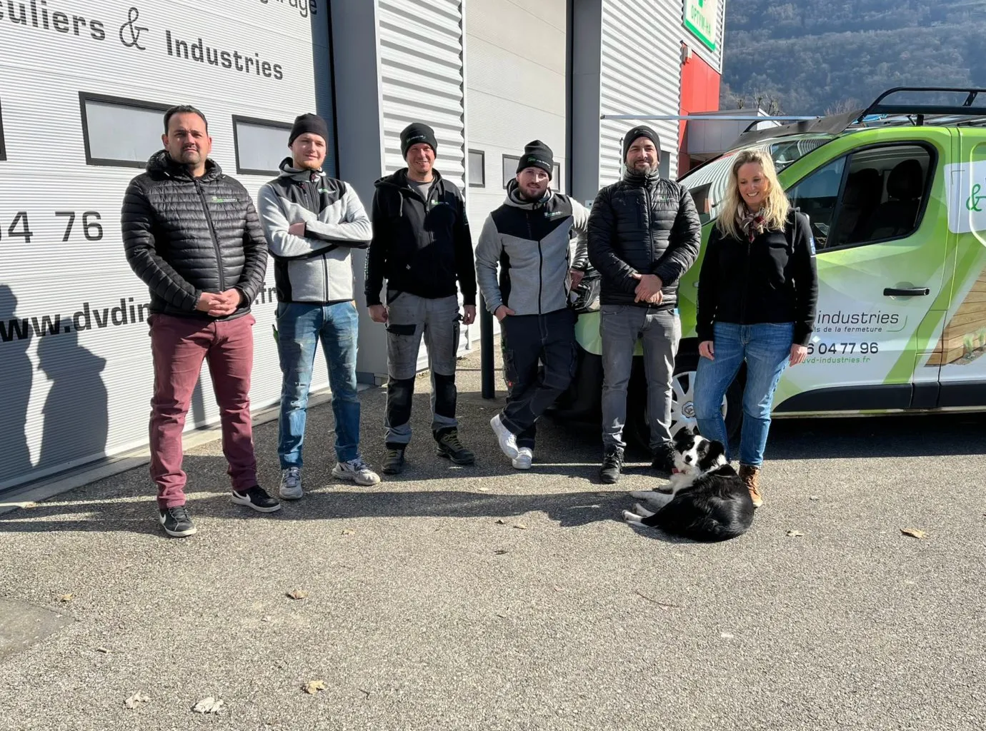 Cinq hommes et une femme debout devant un bâtiment industriel avec une camionnette verte et un chien assis.