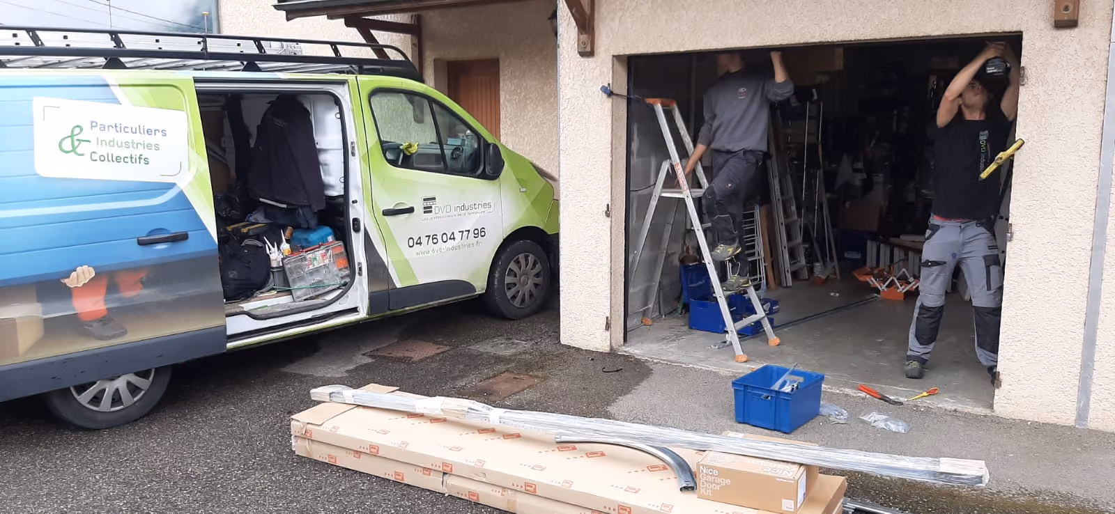Deux ouvriers installent une porte de garage, camion DVD Industries garé avec outils et matériaux.
