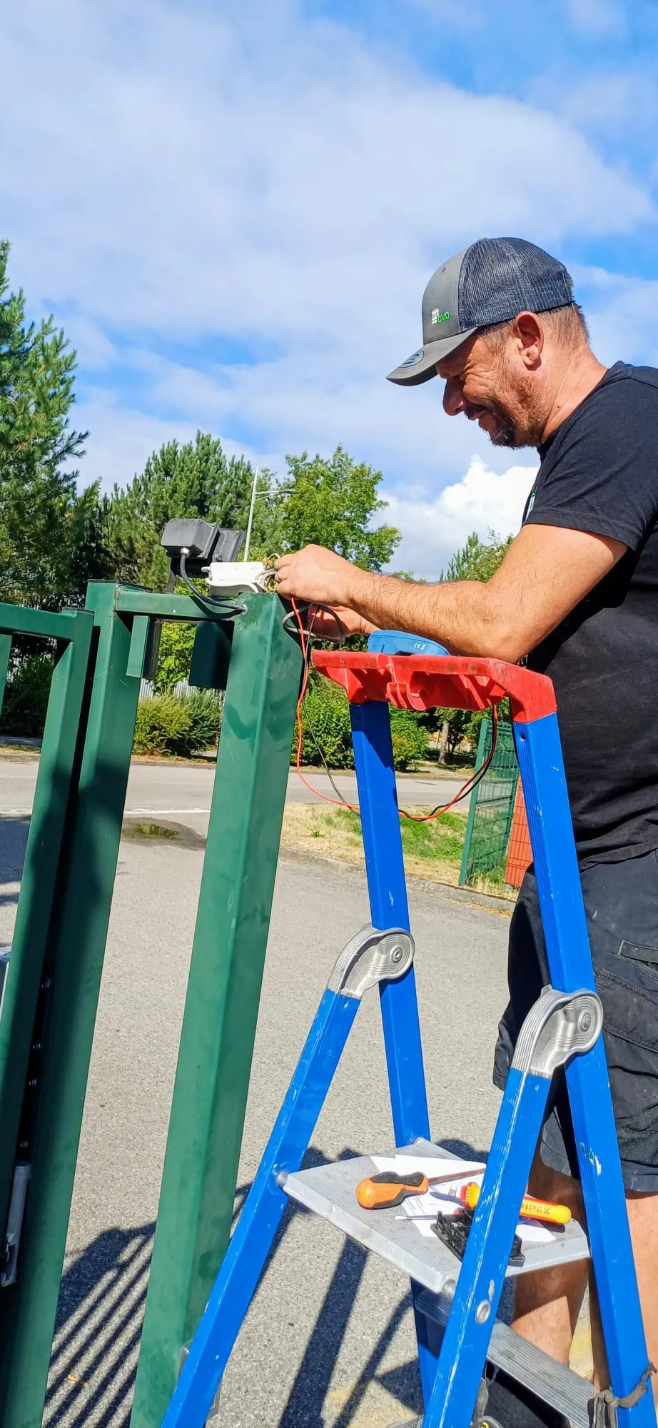 Homme sur une échelle bleue travaillant sur un dispositif électrique fixé à une grille verte extérieure.