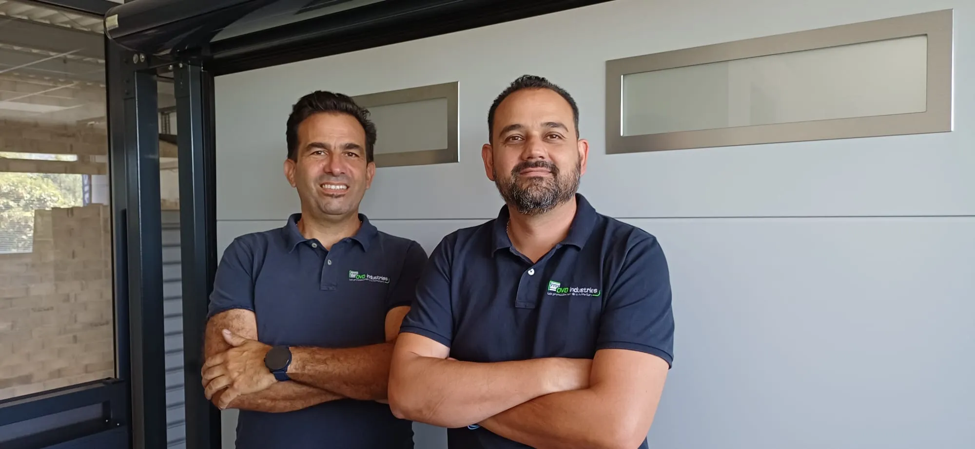 Deux hommes souriants en polo bleu marine avec logo d'entreprise, bras croisés, debout côte à côte.