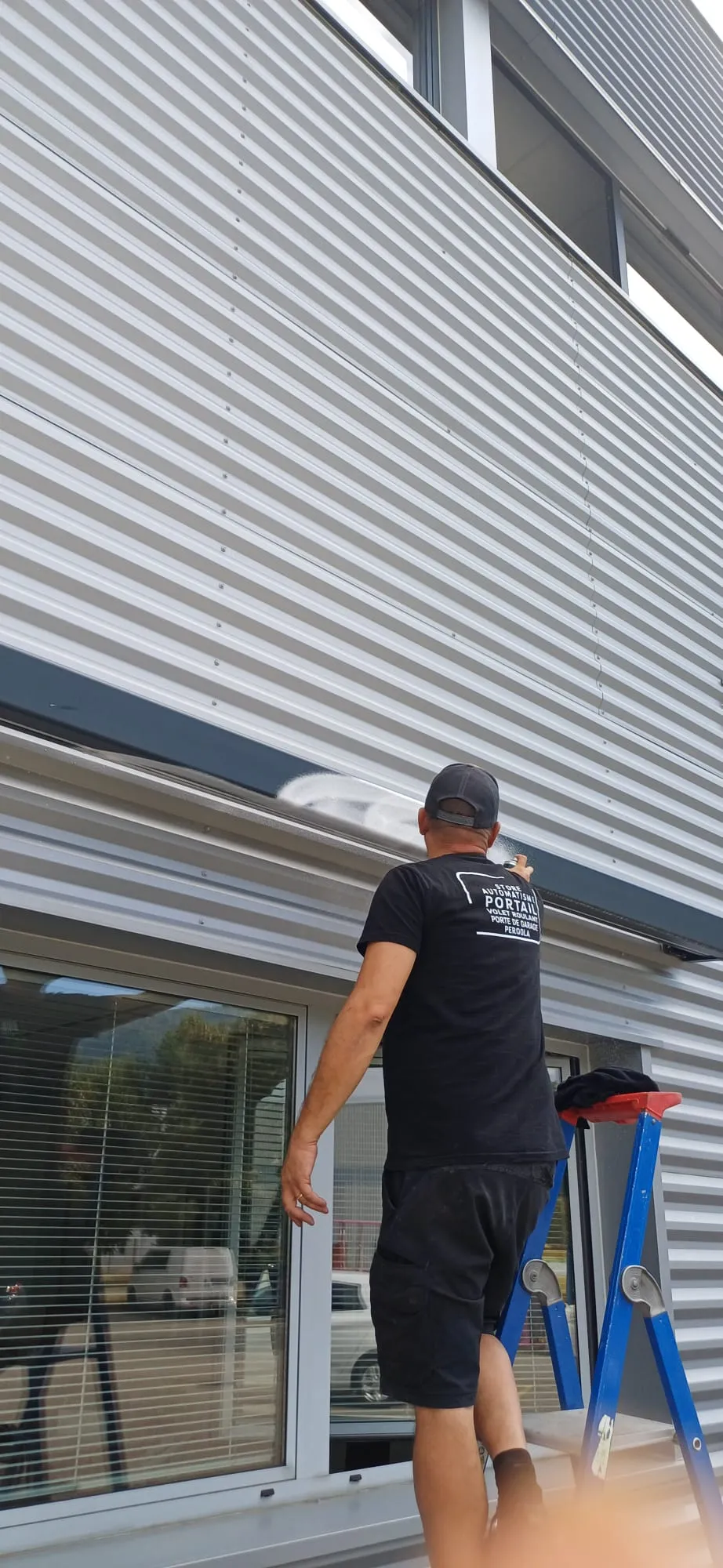 Un homme debout sur une échelle bleue nettoie la façade métallique d'un bâtiment moderne.