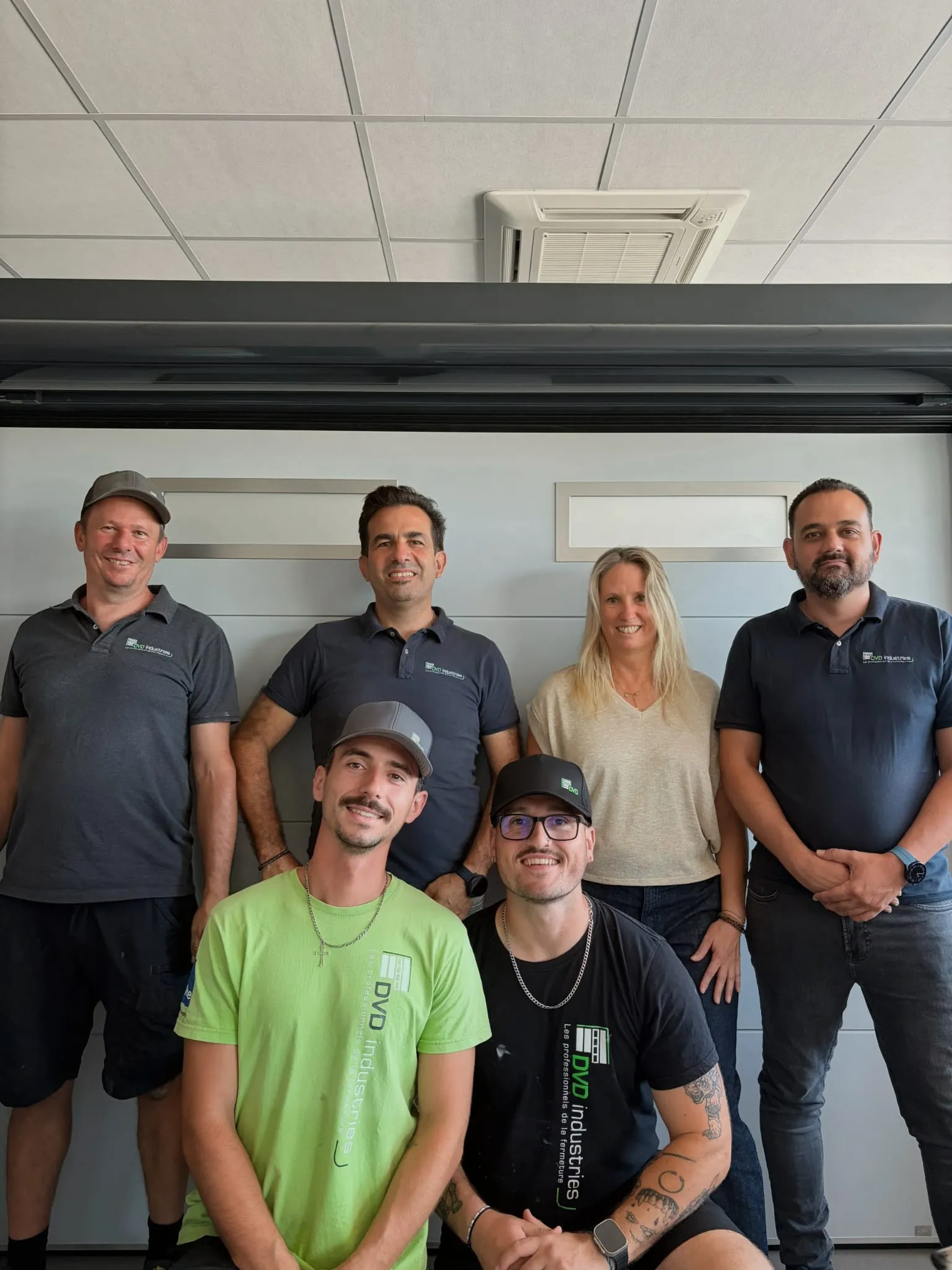 Groupe de six personnes posant devant un mur gris, cinq hommes et une femme souriants en tenue décontractée.