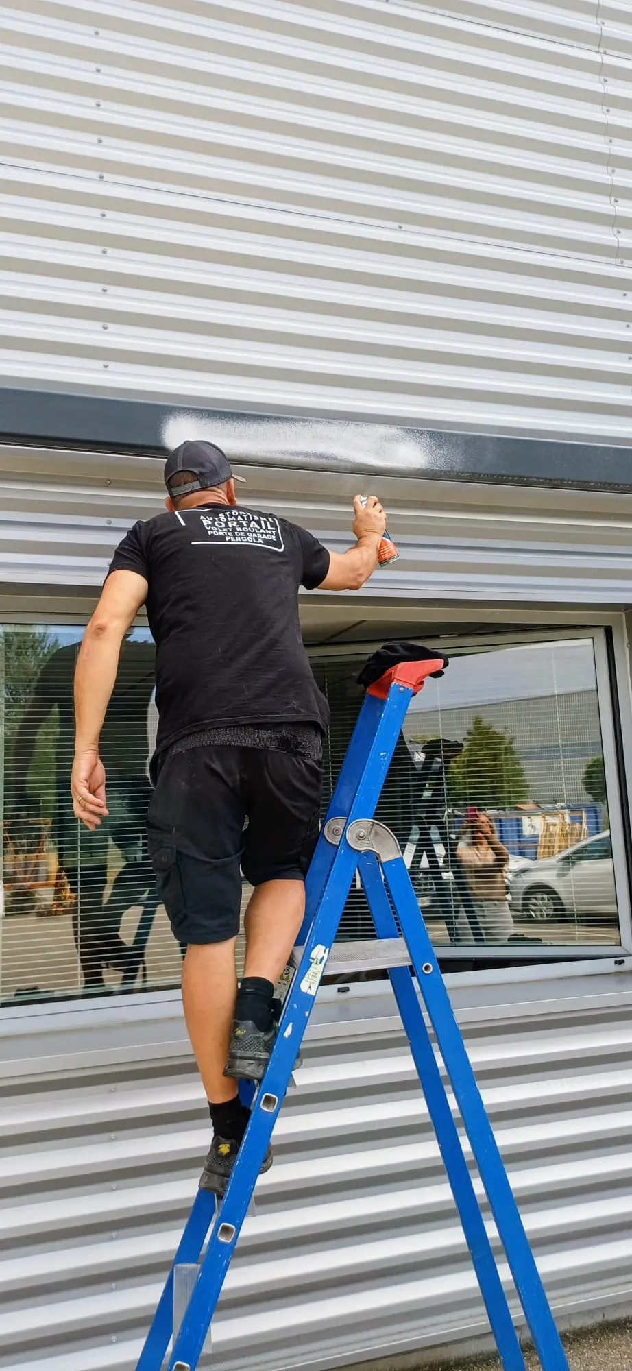 Un homme debout sur une échelle bleue, en train de pulvériser de la peinture blanche sur une façade métallique.
