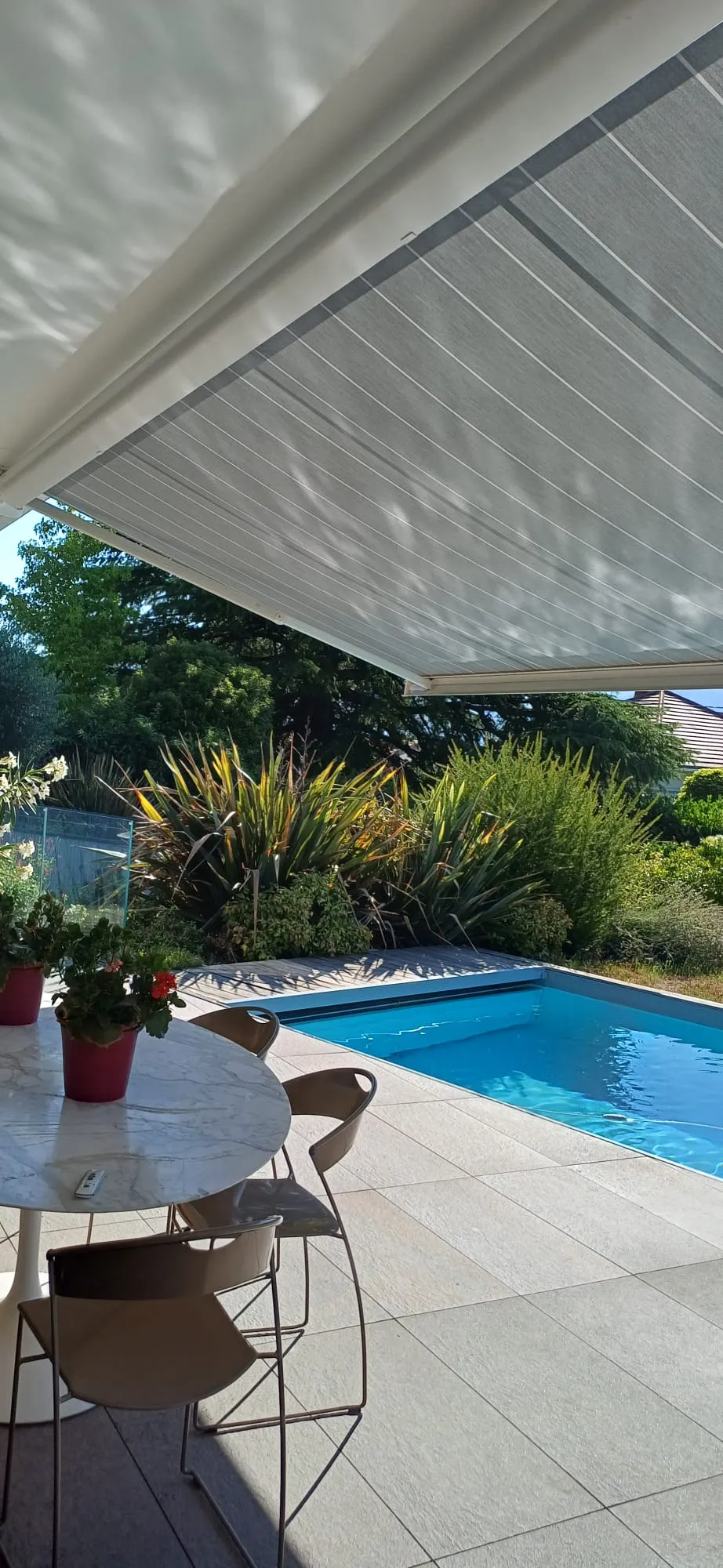 Terrasse extérieure avec table ronde, chaises, plantes en pots, piscine et auvent blanc.