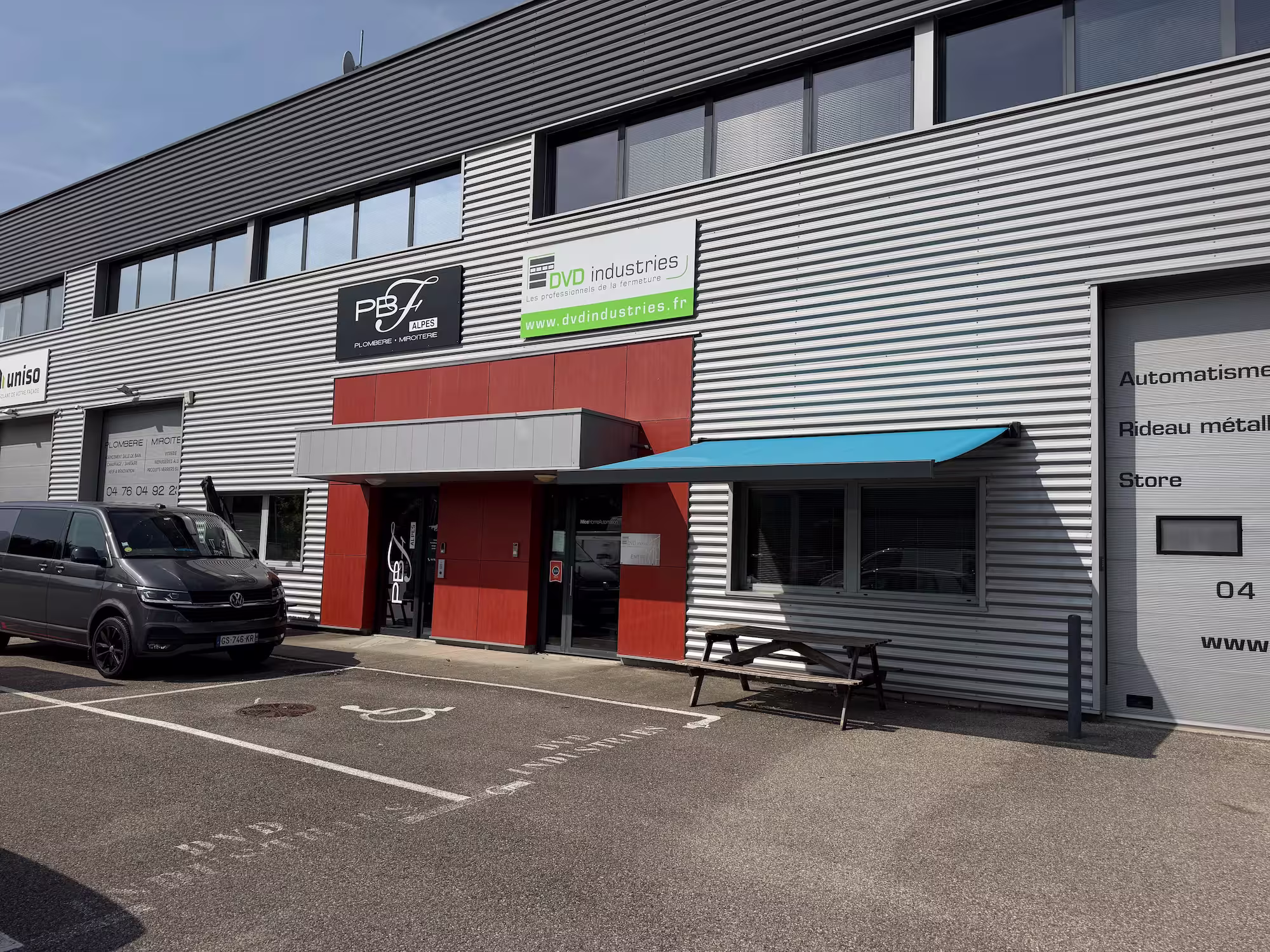 Façade industrielle avec entrée rouge, auvent bleu, van noir garé et banc en bois.