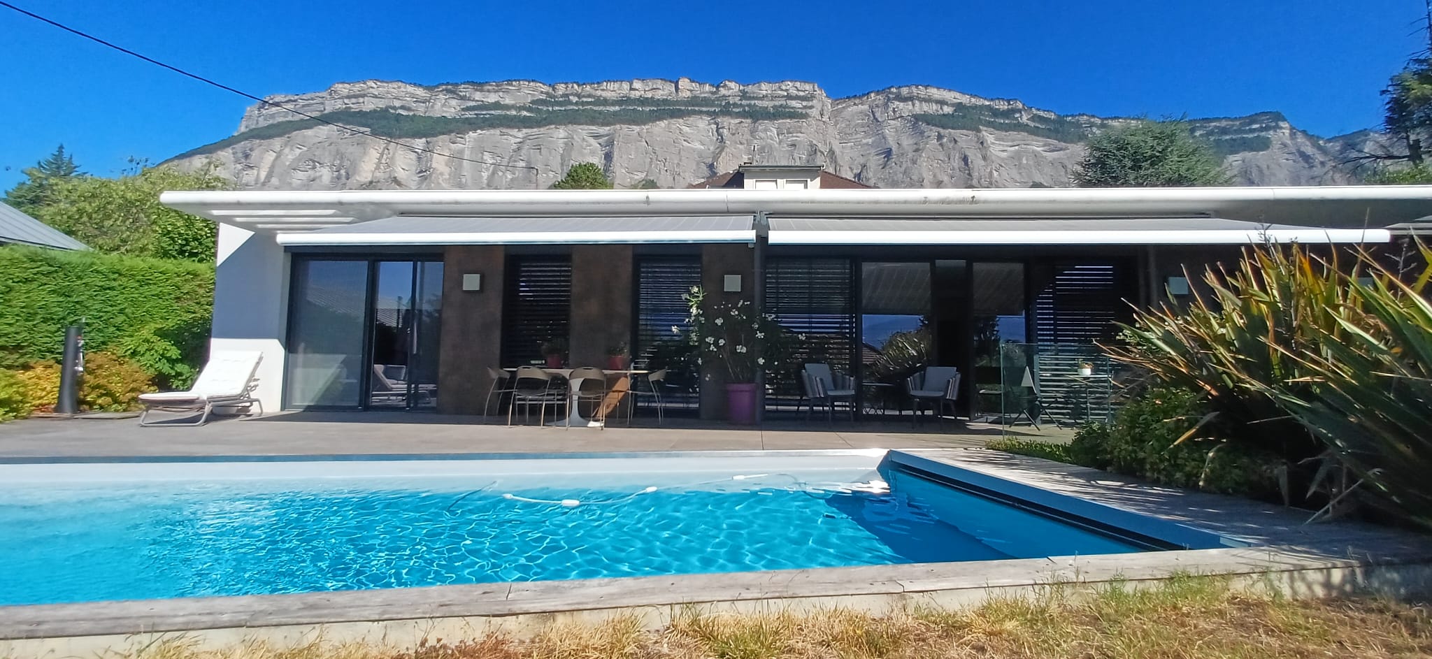 Piscine bleue devant une maison moderne avec montagne rocheuse en arrière-plan sous ciel bleu.