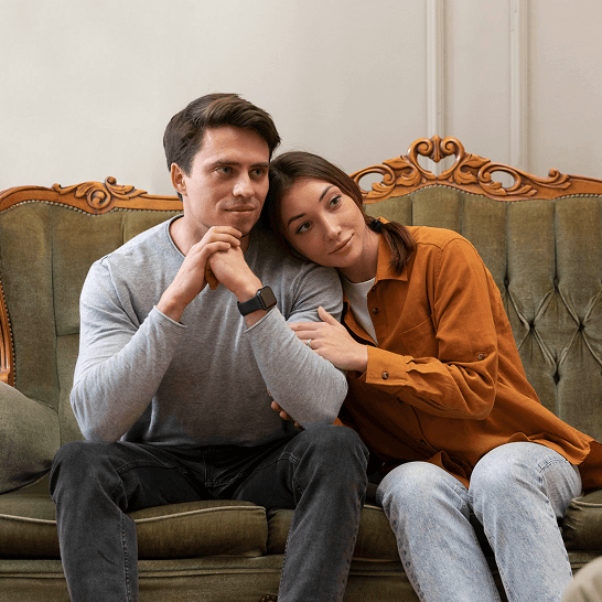 Man and woman sitting closely on a vintage couch, the woman resting her head on the man's shoulder.
