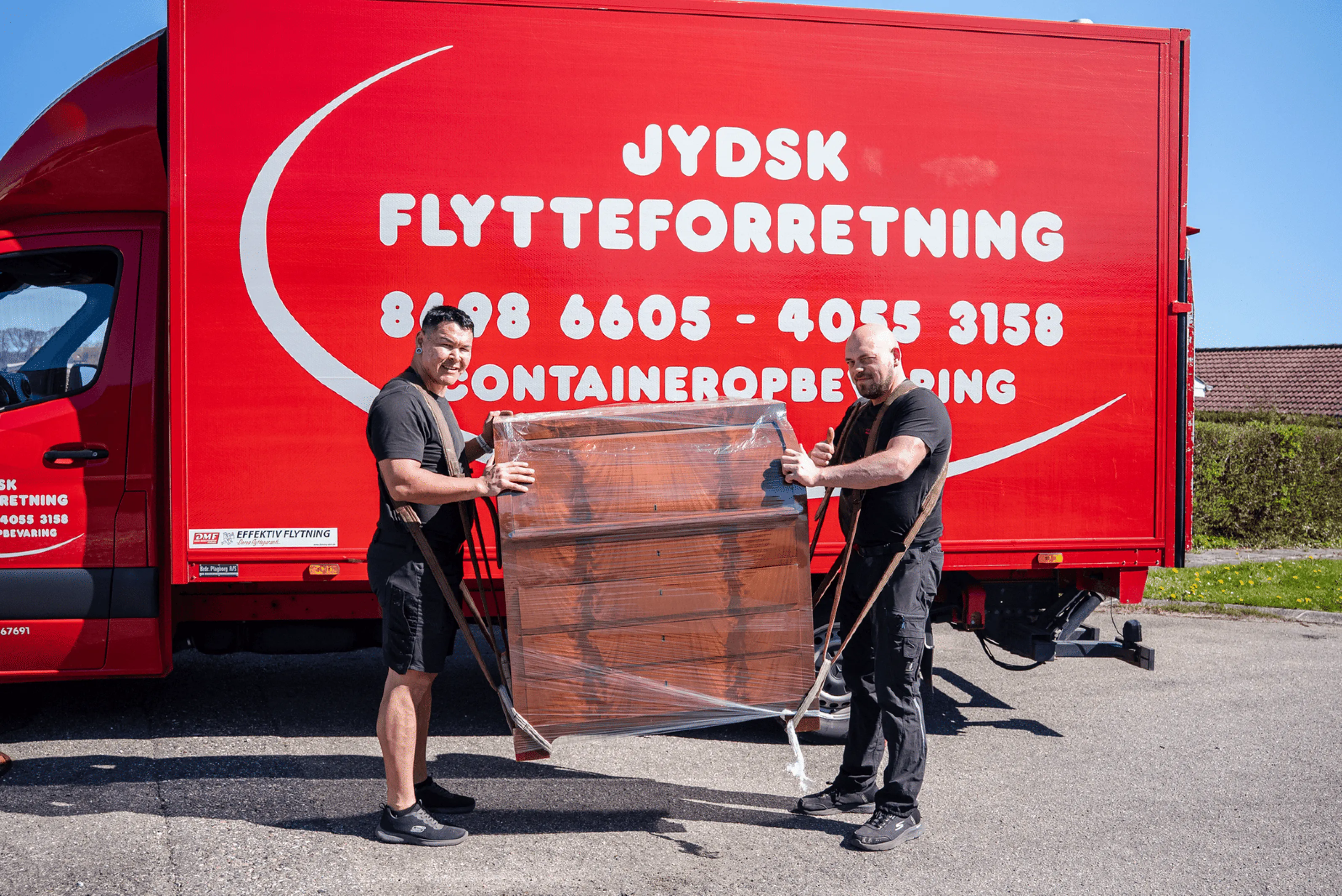 Two movers carrying a wrapped wooden dresser in front of a red moving truck with company text.