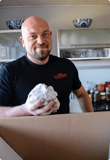 Smiling bald man unpacking a box in a kitchen, holding crumpled paper for packing.