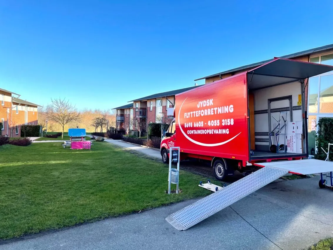 Red moving truck with open back and ramp parked near apartment buildings on green lawn.
