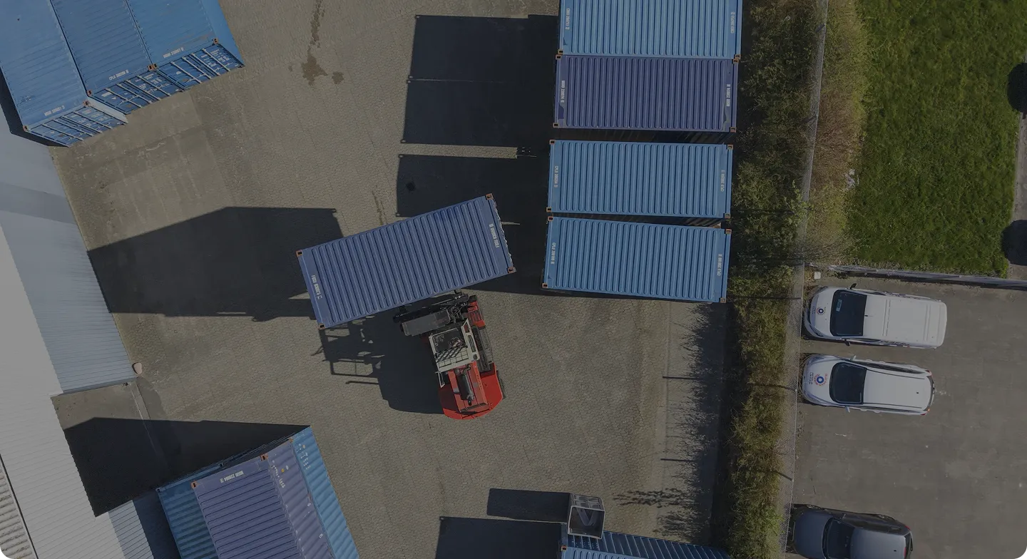 Aerial view of a forklift lifting a blue shipping container in a container yard with parked vehicles nearby.