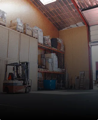 Industrial warehouse interior with stacked pallets, shelving, a forklift, and a partially open overhead door.