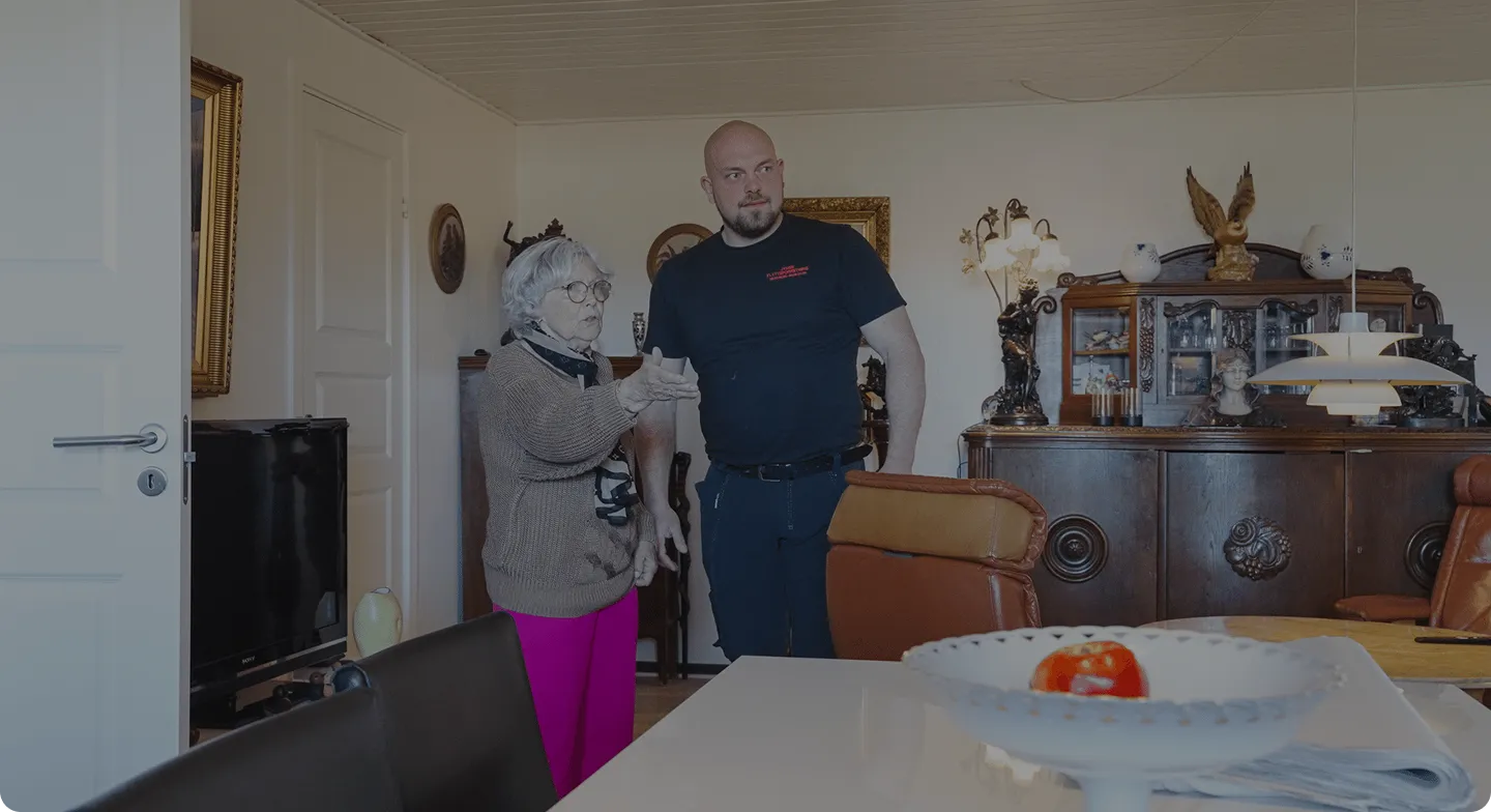 Elderly woman in pink pants gestures while talking to a standing man in a living room with vintage furniture.