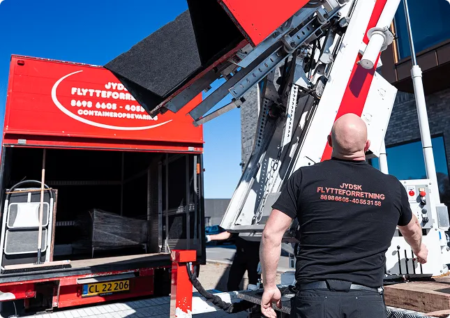 Worker operating a mechanical lift to move items into a red moving truck under blue sky.