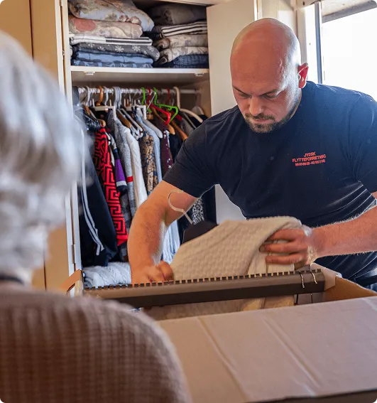 Man folding a blanket into a box while standing by an open wardrobe filled with clothes.