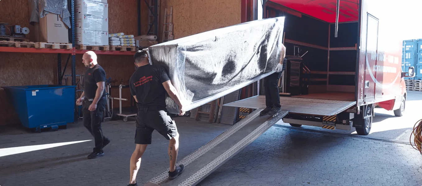 Two movers carrying a wrapped couch up a ramp into a red moving truck with a third man standing nearby.