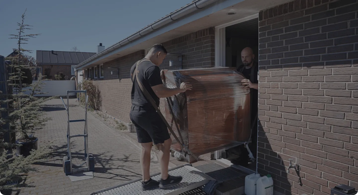 Two men moving a large wrapped wooden cabinet into a brick house through an open door.