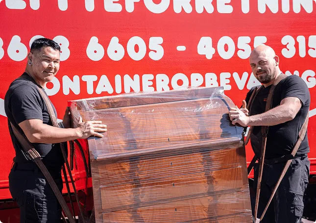 Two men in black shirts carry a wrapped wooden dresser in front of a red wall with white text.
